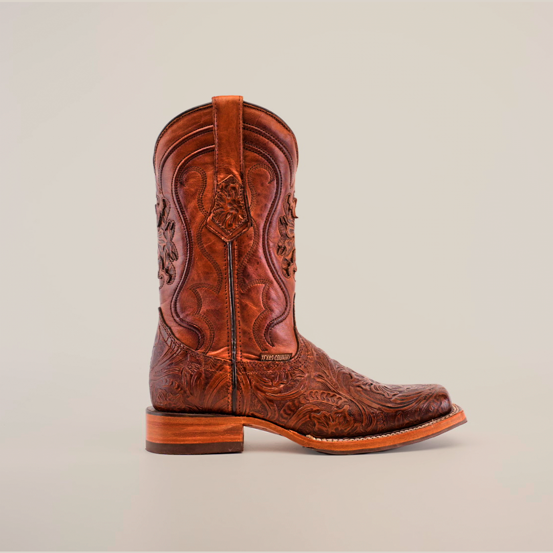 The Handtooled Leave Brown cowboy boot, crafted from premium cowhide, showcases intricate patterns and embroidery. Featuring a rodeo toe and slip-resistant sole, the boot is positioned against a plain background with a convenient pull strap at the top.