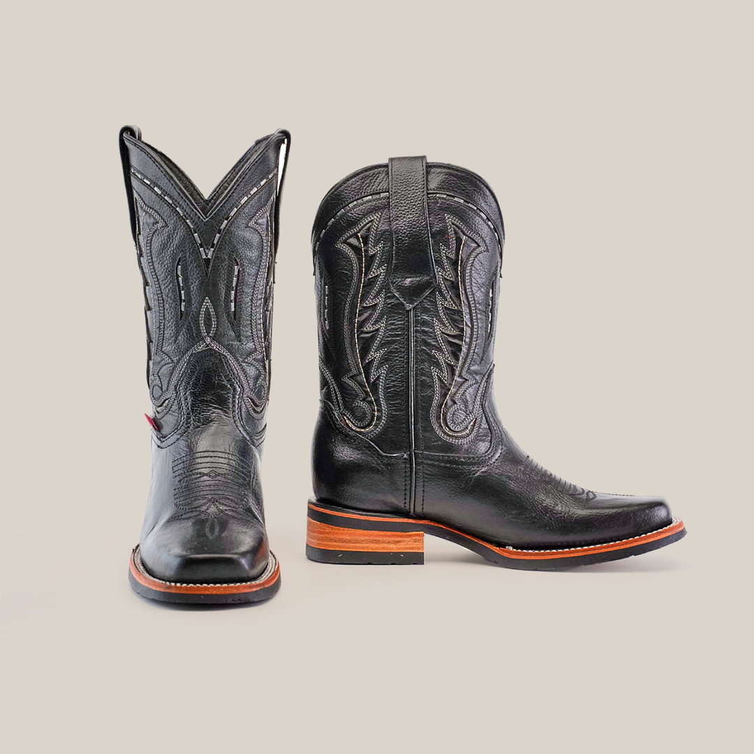 The Barcelona Black Slip Resistant Sole cowboy boots, handcrafted from premium cowhide, showcase intricate stitching. Featuring a high shaft with pull tabs and a brown wooden heel and sole, one boot is shown front-facing and the other side-facing on a white backdrop.