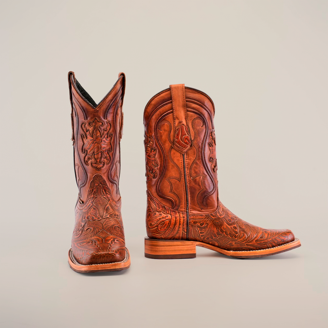 The Handtooled Leave Cogñac boots, featuring a Rodeo Toe and slip-resistant sole, are elegantly displayed in rich brown cowhide leather with intricate embossed patterns; one boot stands upright while the other lies on its side against a pristine white background.