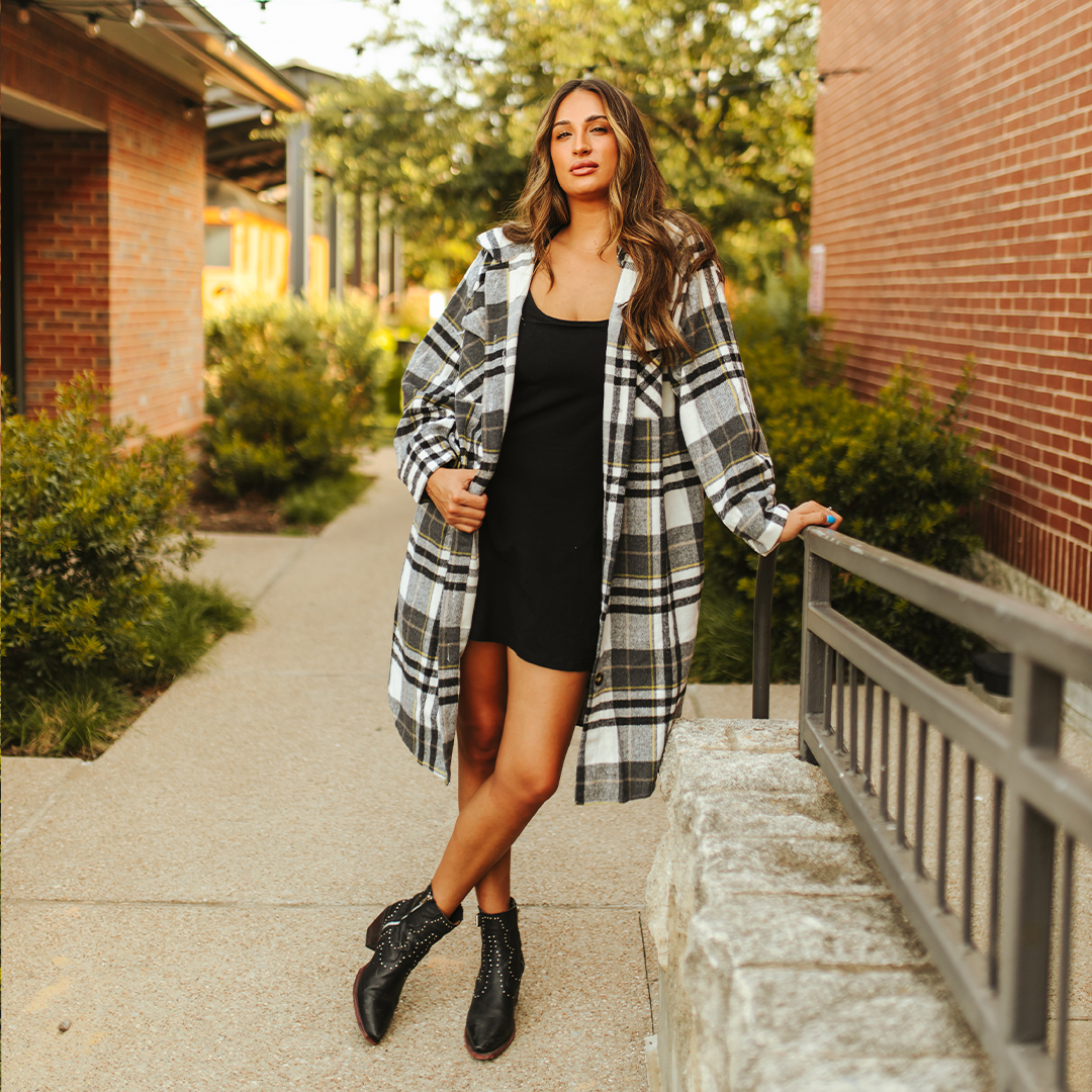 Wearing the LB Black Plaid Fleece Long Button Down JK223-BLK, a woman leans against a stone wall on a sunny day. She stands between greenery and a brick building, one foot forward, hands in her pocket, under a clear sky.