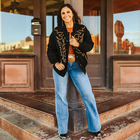 A woman smiles outdoors, exuding edgy elegance in an LB Black Western Stitch Bomber JO241-BLK over a brown top, wide-leg blue jeans, and sandals. Her hands rest on her hips against a backdrop of wooden architecture and a stop sign.