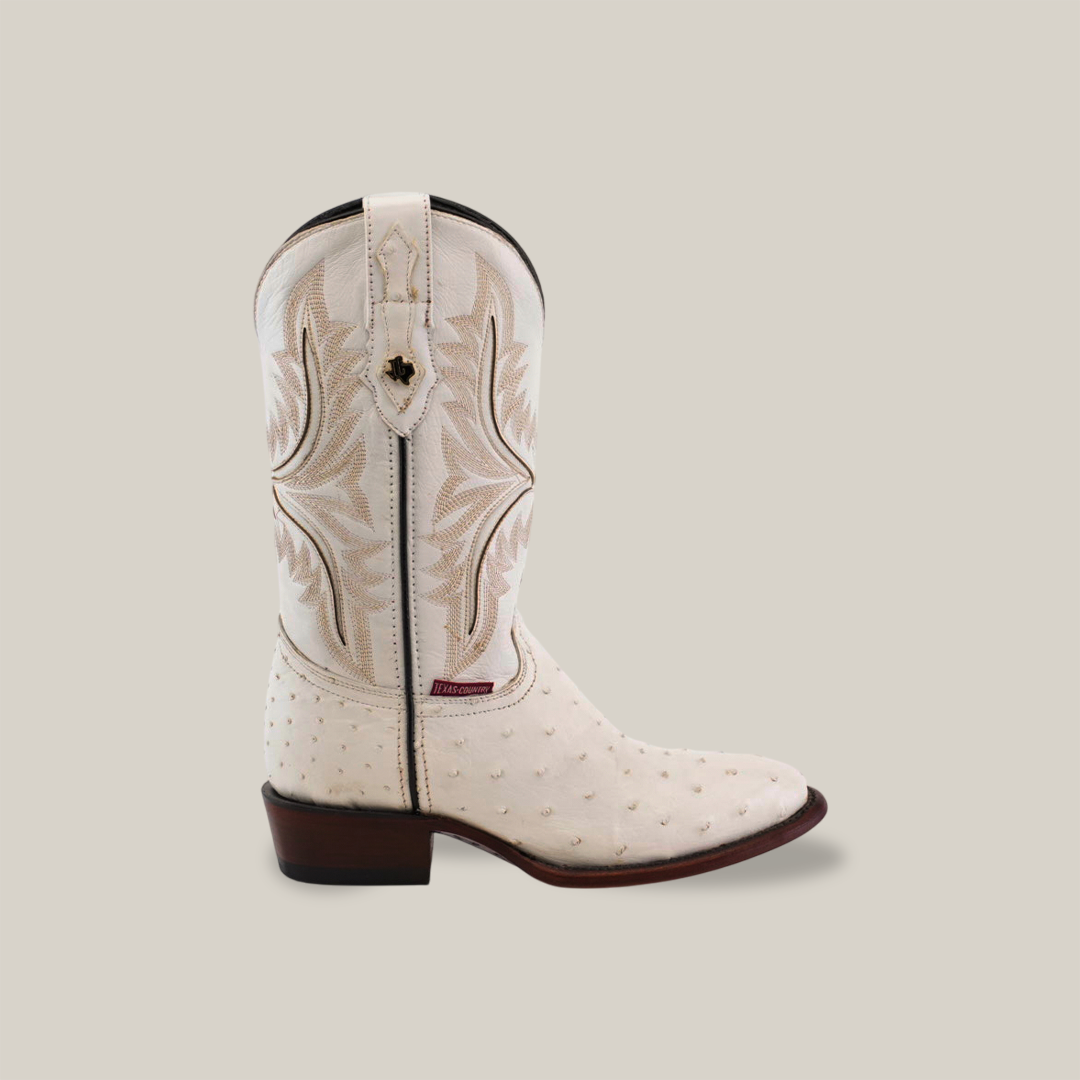 The Exotic Full Quilt Ostrich boot in white features detailed embroidery on genuine ostrich leather. This handmade boot includes a round toe, mid-calf height, brown stitching on a beige background, and a dark brown wooden heel.