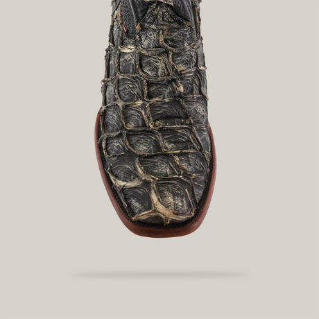 A close-up of an Exotic Piraruco Fish boot, Rustic Bone color, with a black and brown embossed alligator pattern. Crafted with a narrow square toe and smooth light brown sole, set against a plain light gray background.