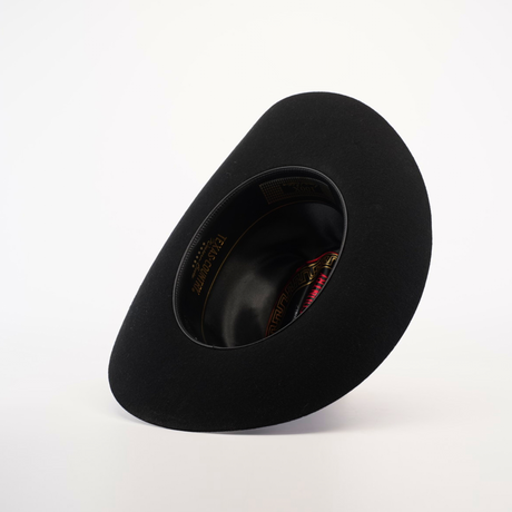 The X 2 Carnales Black, a classic wide-brimmed felt hat, rests upside down on a plain white background. Its inner lining is slightly visible, showing hints of gold text and red fabric.