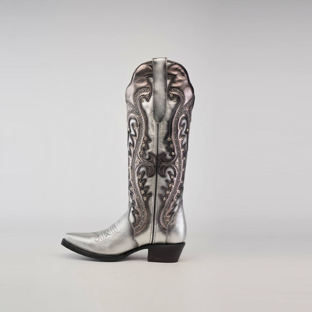 The Valentina Silver Tall Shaft boot, with a snip toe, is a stylish cowboy boot crafted from premium leather. It features metallic silver tones, elaborate embroidery, and ornate stitching with a mid-calf height and black heel against a plain light background.