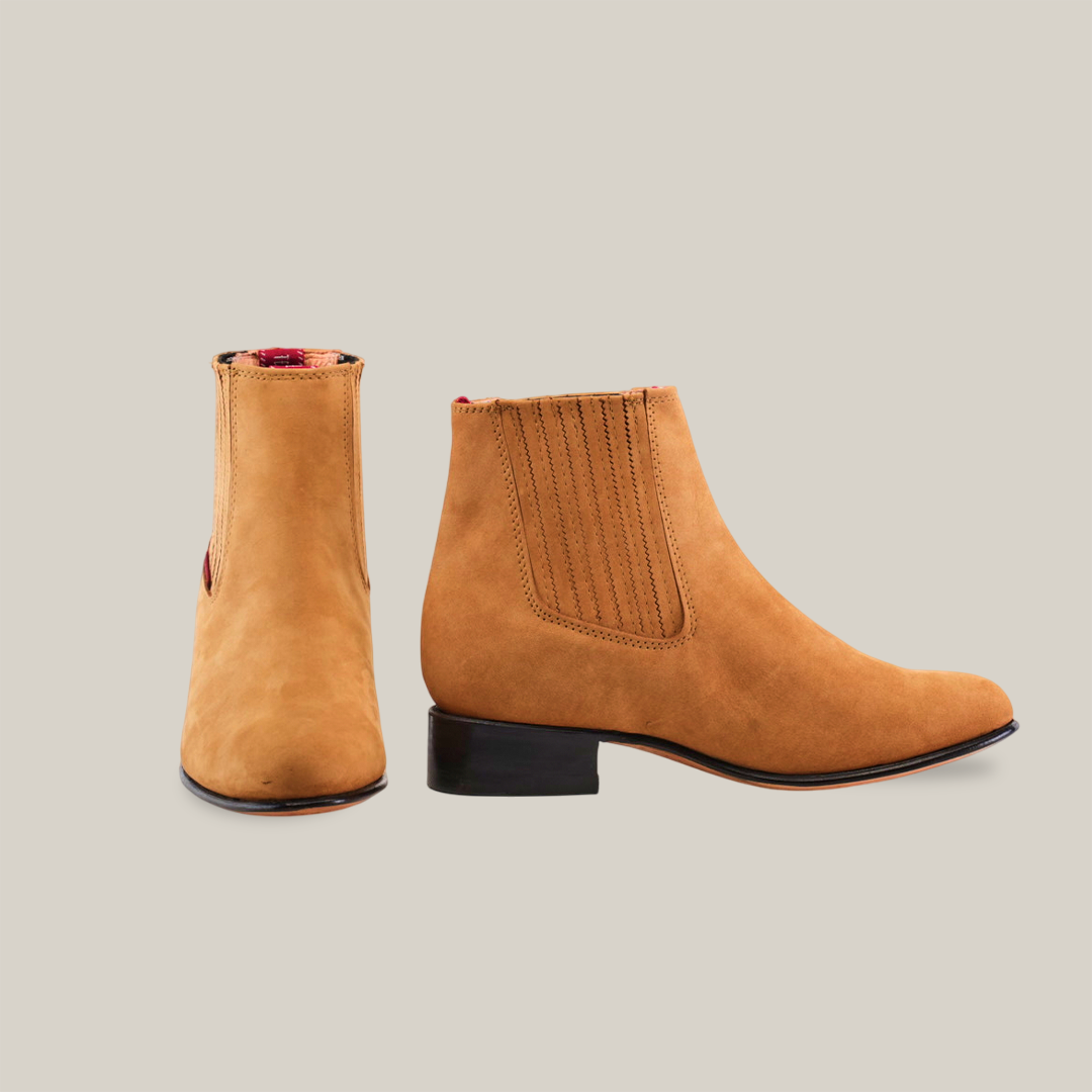 The Prime Suede Honey Round Toe ankle boots, handcrafted in light brown honey suede with elastic side panels and low black heels, are elegantly displayed on a white background. One boot faces forward while the other is shown from the side, showcasing their Western-inspired elegance.