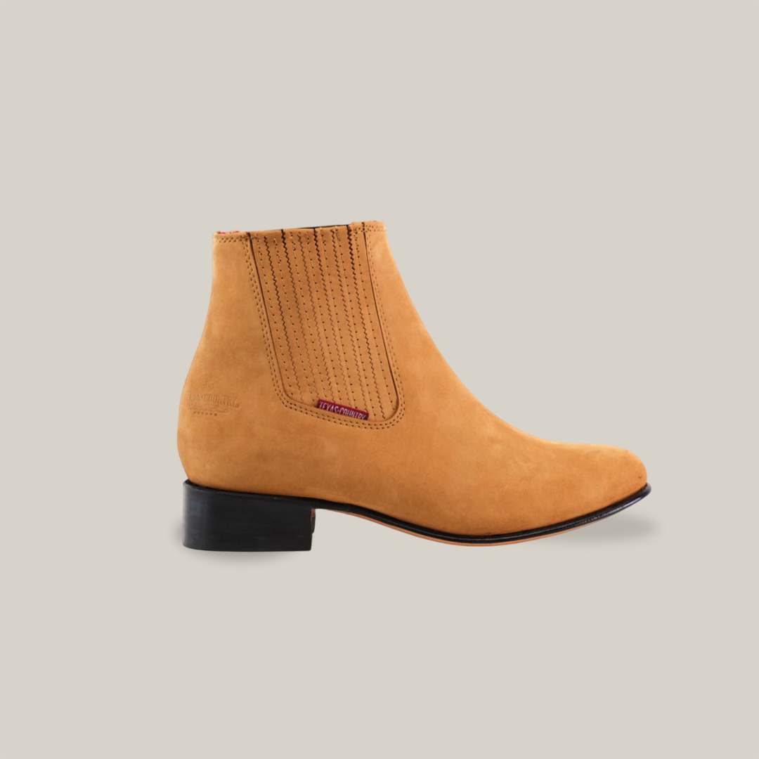 The Prime Suede Honey Round Toe is a sophisticated honey suede Chelsea boot with a low black heel and Western flair. It boasts elastic side panels with vertical stitching, a purple side label, and handcrafted elegance. Photographed against a white background, it exudes style.