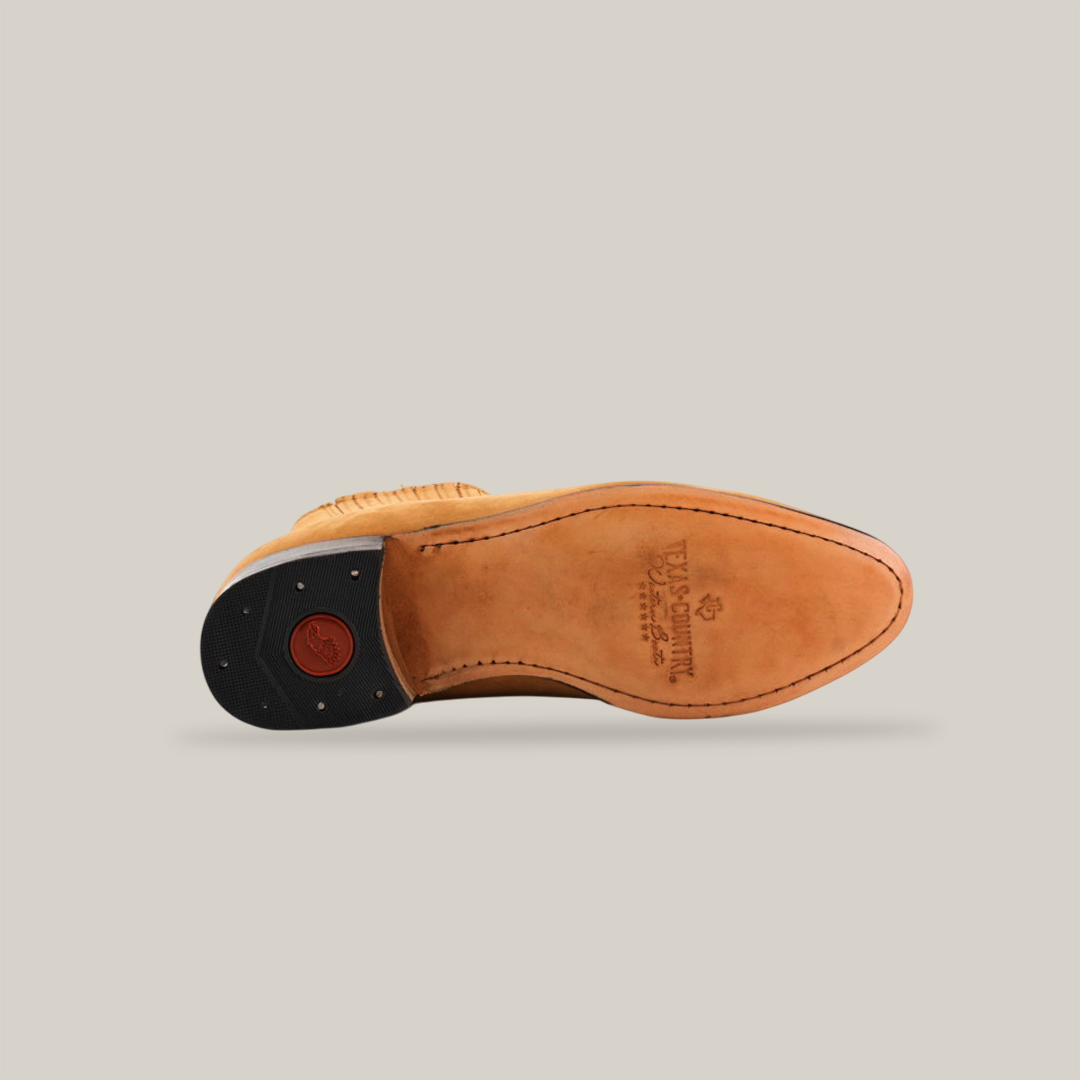 The image depicts the underside of the Prime Suede Honey Round Toe shoe, resembling Western footwear, on its side. The sole displays intricate stitching and an embossed heel logo. A black heel with a red circular insert contrasts with the plain white background.