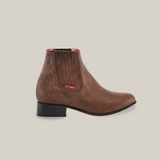 A side view of the Alamo Brown - Round Toe shows a handcrafted brown leather ankle boot with a low black heel, elastic side panels, red label, and textured finish.