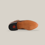 The Alamo Brown - Round Toe is shown sideways, featuring a tan sole with a black heel and a red circular logo. The premium leather is highlighted with stitched detailing along the edges against a plain white background.