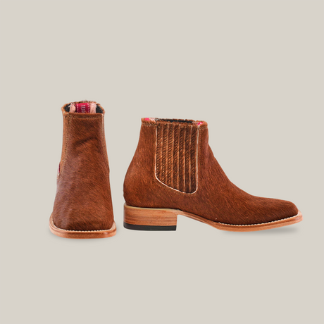 The Cowhide Hair Camel Square Toe boots are displayed, showcasing their distinct brown leather with furry cowhide texture. The right boot is facing forward, while the left is angled to reveal the side view and red and white plaid interior lining.