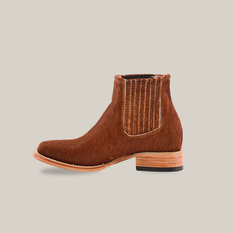 The Cowhide Hair Camel Square Toe is a brown ankle boot featuring a cowhide hair-like finish, pull-on design, vertical elastic side panels, a flat sole with light wood detailing, and a small black heel.