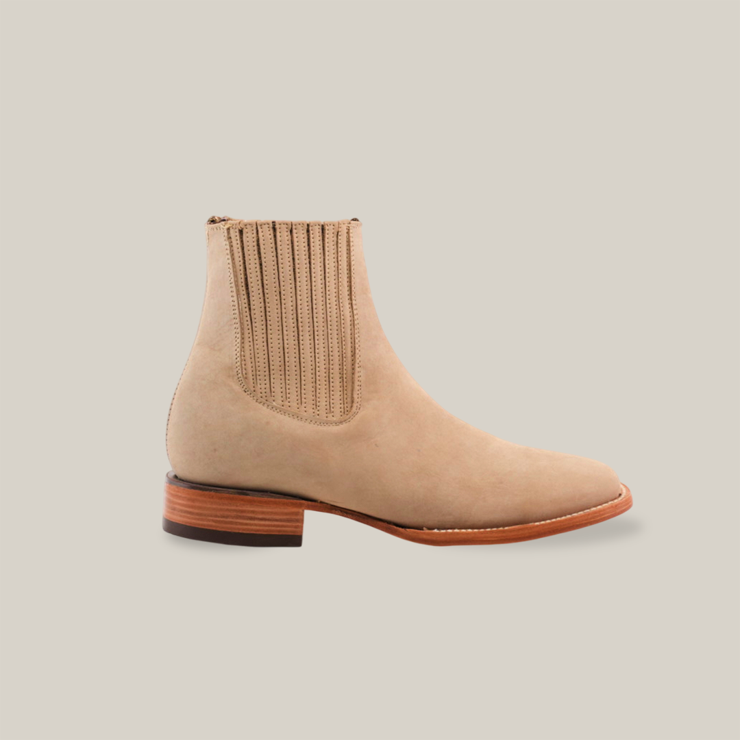 The Prime Suede Pearl Square Toe boot combines beige suede with cowboy flair, featuring side elastic panels with vertical stitching, a square toe, and a wooden sole highlighted by a dark heel. The handcrafted boot is beautifully showcased on a plain white background.