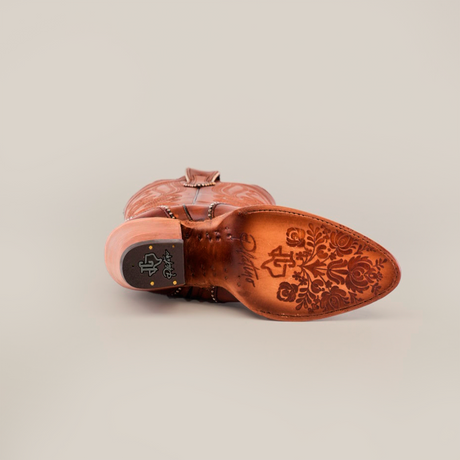 The image shows the underside of a Linda Tall Shaft Tan - J Toe cowboy boot, featuring a premium leather sole with intricate floral engravings and a small black and gold emblem near the heel. The background is plain and light-colored.