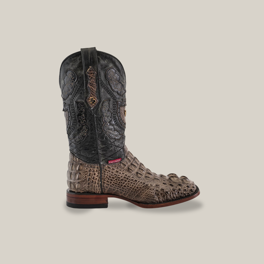 The Exotic American Alligator Head - Gray - Square Toe boot combines handcrafted alligator leather with a brown, textured crocodile lower and intricately-patterned black upper. It features a brown sole and side pull strap, showcased against a plain light gray background.