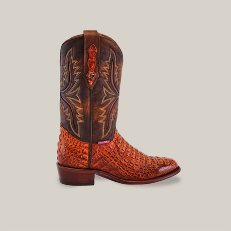 This side view of the Exotic Caiman Hornback - Brandy - Round Toe cowboy boot showcases its authentic caiman leather with a textured red-brown lower and dark brown upper, adorned with intricate tan stitching. It features a low heel and pull strap, embodying rugged elegance.