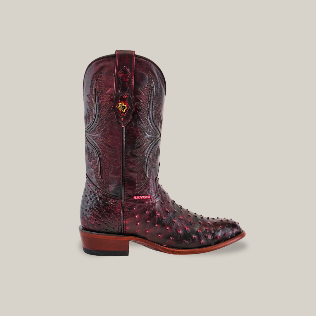 The Exotic Full Quilt Ostrich - Black Cherry - Round Toe boot showcases intricate stitching, a decorative side pull tab, and a combination of a smooth upper with full quilt ostrich texture below, elegantly resting on a wooden heel.