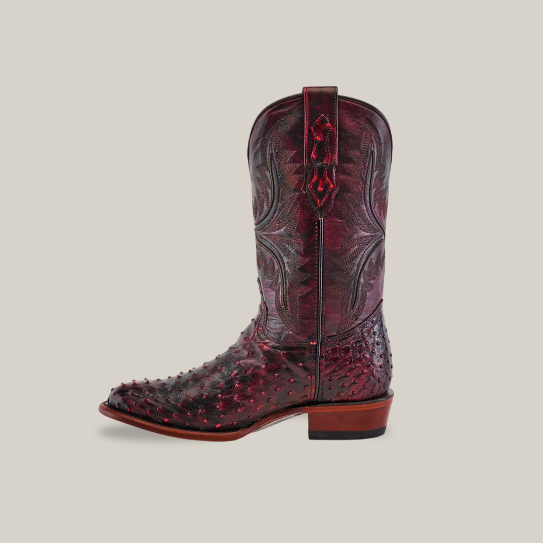 A single handcrafted Exotic Full Quilt Ostrich boot in Black Cherry features a glossy finish, textured ostrich pattern on the lower part, and intricately stitched designs on the upper. It has a brown sole with a slight heel set against an off-white background.