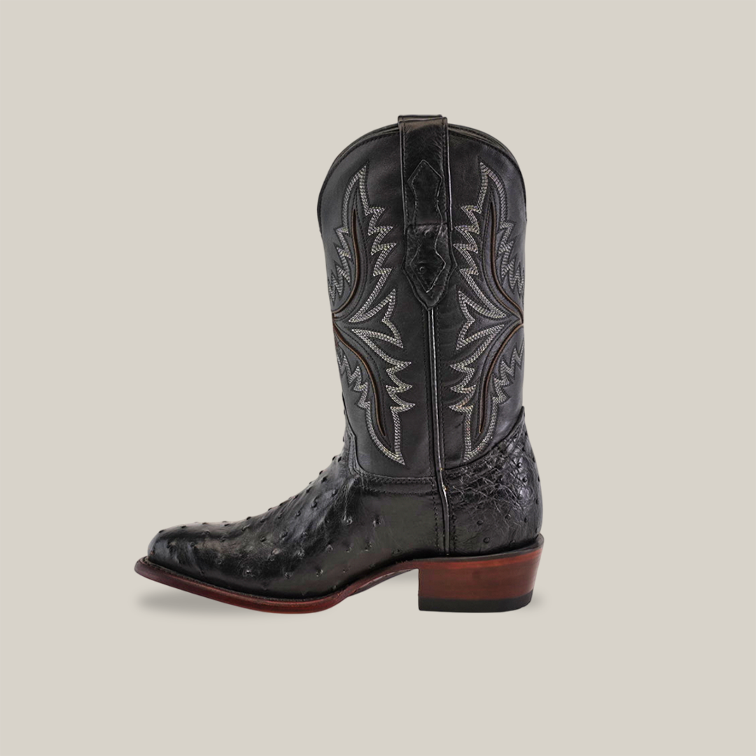 The Exotic Full Quilt Ostrich in black features decorative quilt-like stitching and a textured finish. This Western single boot showcases a round toe and a wooden heel, all presented against a plain, light brown background.