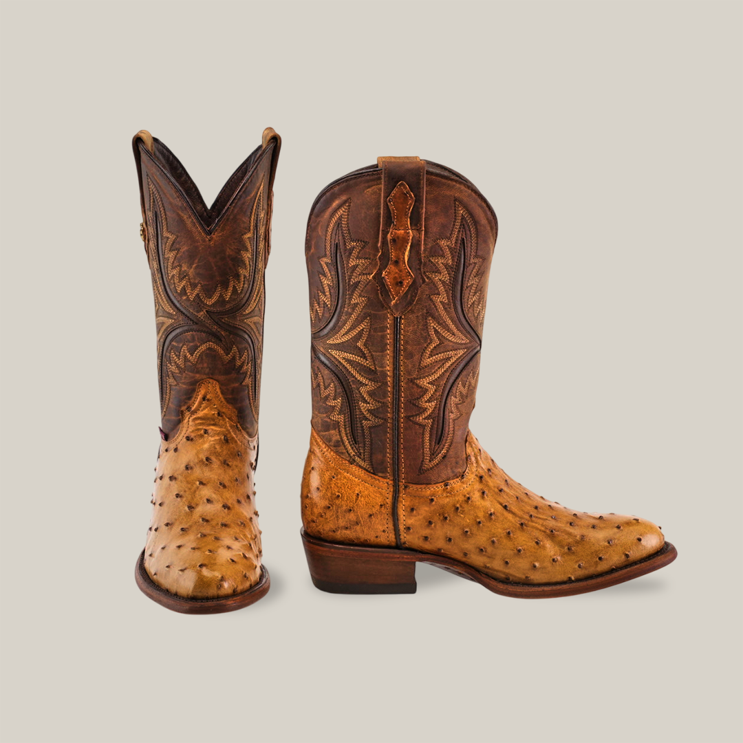 The Exotic Full Quilt Ostrich - Paja boots showcase Western heritage with tan ostrich leather on the foot and a dark brown shaft featuring intricate stitching. One boot stands upright, while the other is angled forward against a light background.