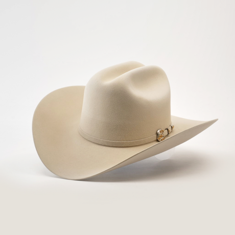 The 1000X Imperial Belly hat is a beige cowboy style with a wide brim, adorned with a decorative band featuring a small metallic emblem, set against a plain white background.