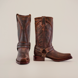 The Azkar Tan boots, crafted from rich cowhide leather, stand side by side on a white background. They feature decorative stitching, a harness with metal rings and studs, pointed toes, and slip-resistant soles for safety—all in a stylish design.