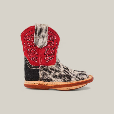 The Cowhide Hair - Red Suede Shaft - Square Toe childrens cowboy boot showcases a red suede shaft with embroidery, a black and white cowhide patterned lower part, and a brown leather sole with a square toe. It is set against a plain, light beige background.