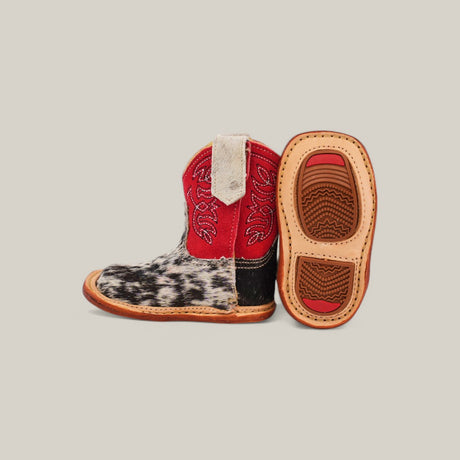 The Cowhide Hair - Red Suede Shaft - Square Toe baby boots feature cowhide fabric and red suede shafts with intricate red stitching. One boot is upright, revealing a charming rustic style, while the other lies on its side to show a textured tan sole against a neutral gray background.