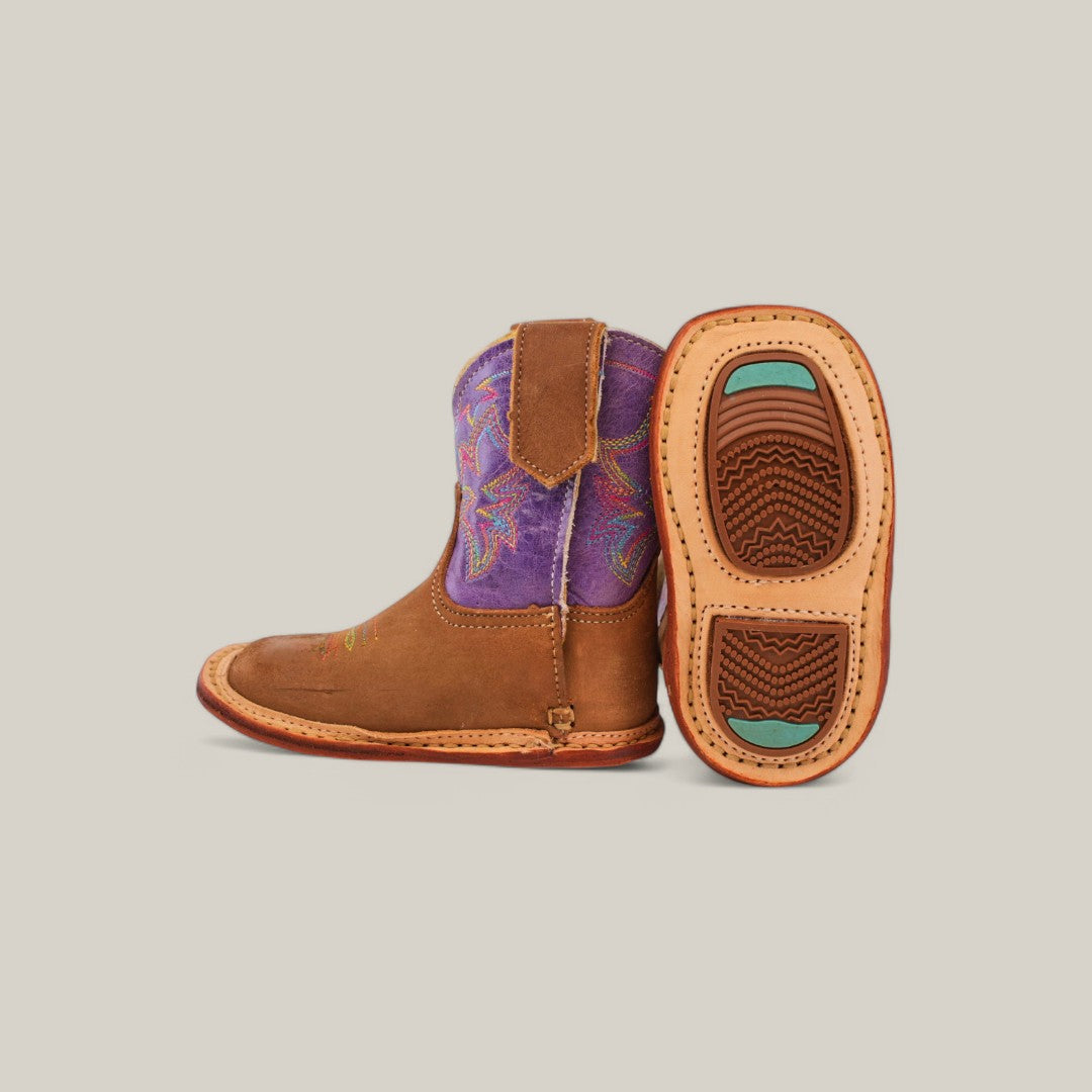 A pair of childrens Lilly boots, one standing upright and the other showing the sole. These cowboy boots feature a camel suede body with violet shafts, colorful stitching, a square toe, and a brown sole with green accents for a playful touch.