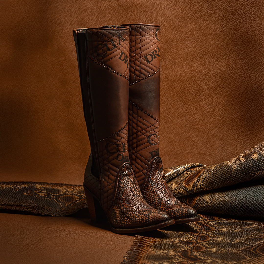 Displayed against rich brown fabric, the Glazed Chestnut Python Laser & Embroidery & Woven boots feature genuine python leather with intricate stitching and a reptile texture. These premium quality boots offer a high heel and decorative patterns for a luxurious and stylish touch.