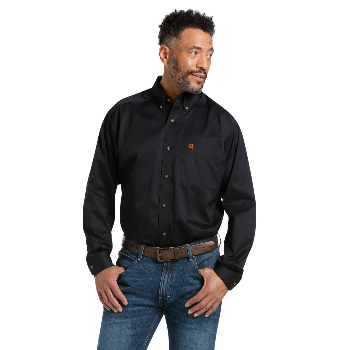 A man in a Solid Twill Classic Fit Shirt Black - 10000502, featuring embroidered chest pocket details, pairs it with blue jeans and a brown belt. Hands in pockets, he looks to his left.