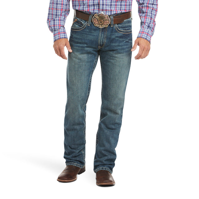 Someone is wearing the M4 Low Rise Boundary Boot Cut Jean in blue, paired with a brown belt featuring an ornate buckle and brown leather shoes. Their plaid shirt is neatly tucked in, highlighting the classic boot cut style. Only the lower half of their body is visible.