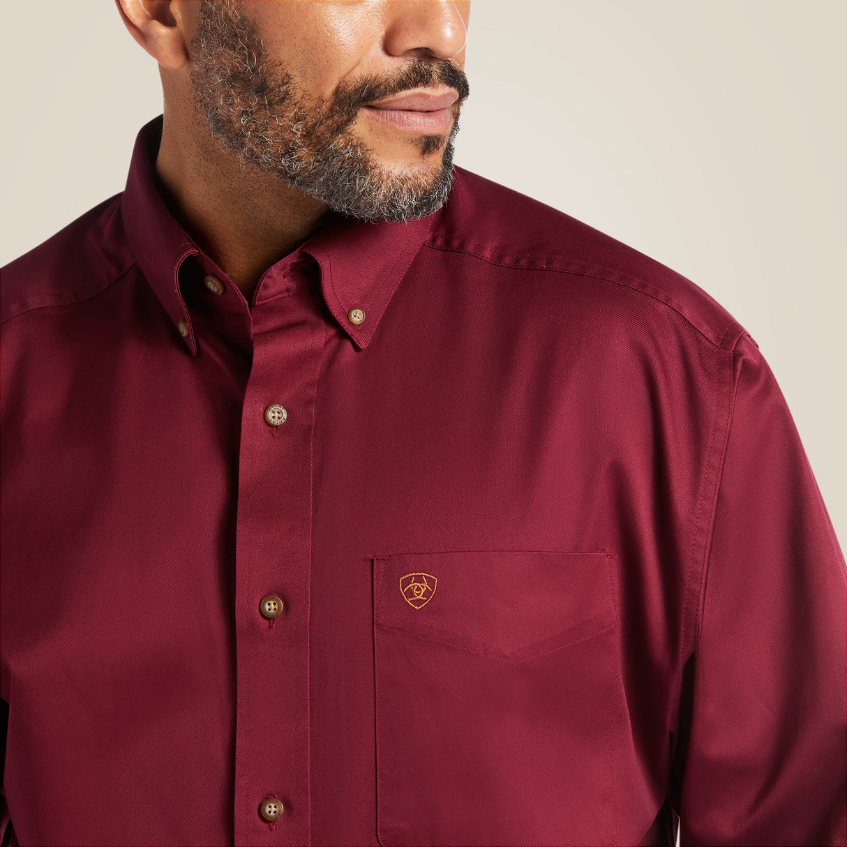 A man in a burgundy Solid Twill Classic Fit Shirt (Product 10012635), featuring a small logo on the chest pocket and buttoned collar, gazes aside with a beard against a neutral background, exuding understated style.