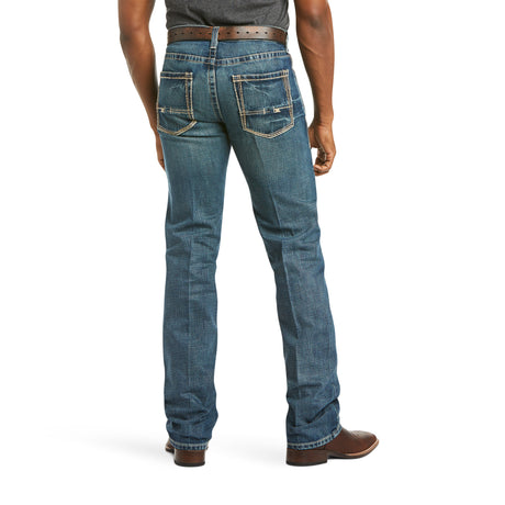 A person stands back to the camera in M5 Slim Boundary Stackable Straight Leg Jeans - Gulch - 10014010 by Ariat, featuring premium cotton blue denim with white stitching. Paired with a brown belt and shoes, two back pockets, boot-cut hems, and a tucked dark shirt for a polished look.