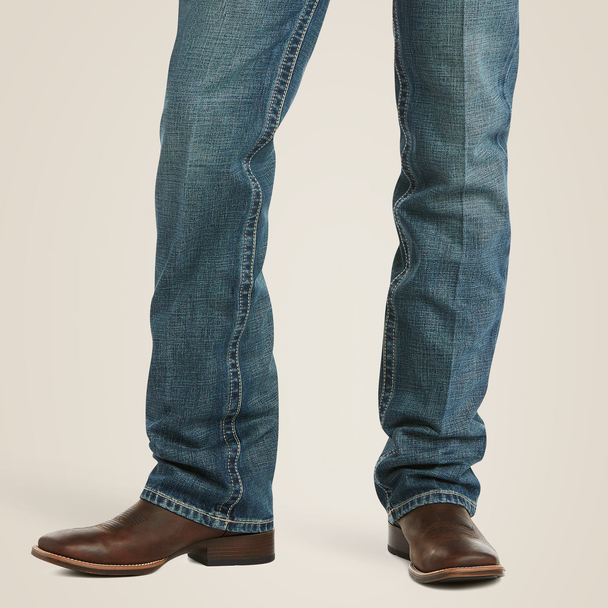 A person in premium M5 Slim Boundary Stackable Straight Leg Jeans - Gulch and polished brown leather cowboy boots stands against a plain background. The jeans have a slightly faded texture and are styled in a straight-leg cut with a low heel on the Ariat boots.