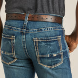 A person models the Ariat M5 Slim Boundary Stackable Straight Leg Jean in Gulch (10014010), featuring visible stitching in blue distressed denim with a brown leather belt. Completing the look is a dark gray shirt for a classic yet modern style.