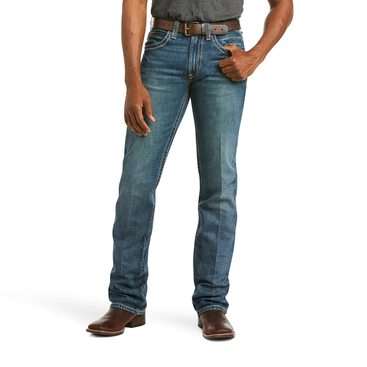 A person is wearing a dark gray t-shirt, Ariat M5 Slim Straight Leg Jeans in blue denim (Gulch - 10014010), and brown leather boots. They have a brown belt with a silver buckle and are standing against a white background with one hand in their pocket.