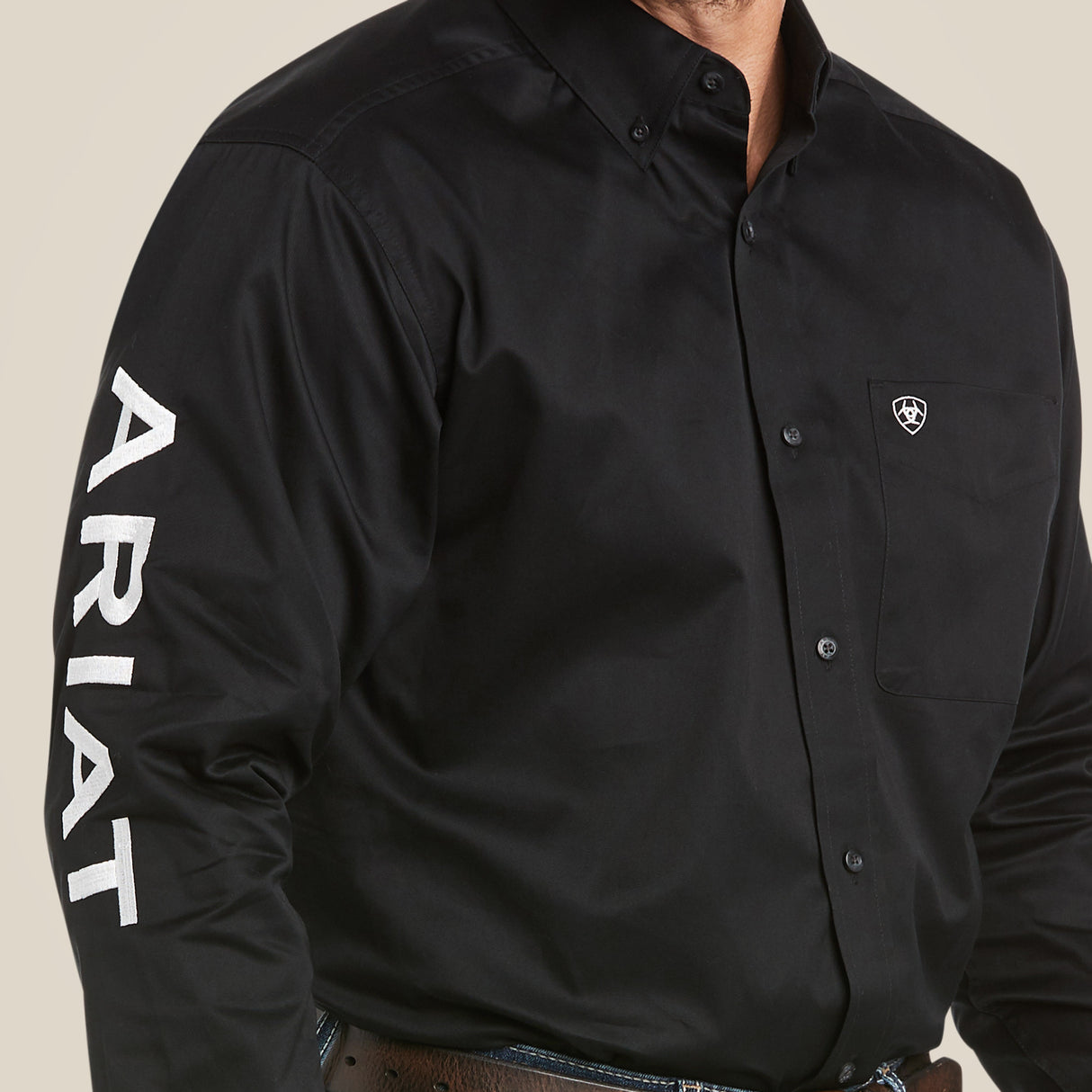 A person wears the Team Logo Twill Classic Fit Shirt Black - 10017497, made of premium cotton with ARIAT in white on the sleeve, a collar, an embroidered shield logo on the chest pocket, and pairs it with a brown belt.