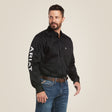 A man with short dark hair and a beard wears the Team Logo Twill Classic Fit Shirt Black - 10017497 featuring embroidered Ariat on the left sleeve, paired with blue jeans. He stands against a beige background, looking off to the side with his right hand slightly raised.