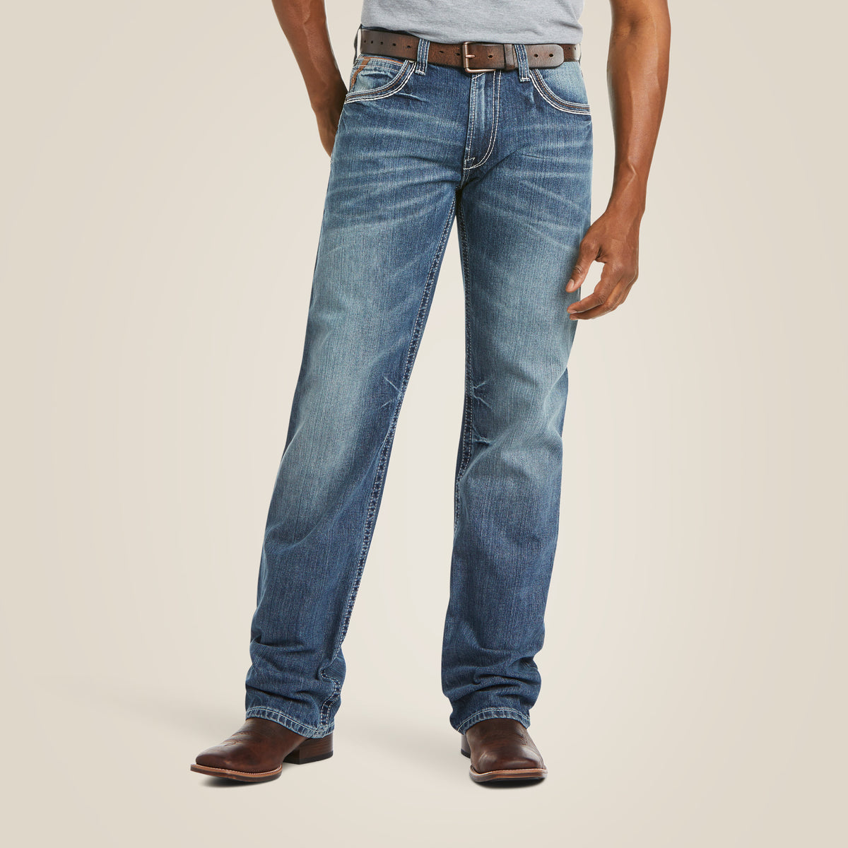 The person is wearing M4 Low Rise Coltrane Boot Cut Jeans - Durango, featuring heavy-duty cotton and light distressing on the thighs. Paired with a gray shirt, brown belt, and brown leather shoes, the upper body is partially hidden.