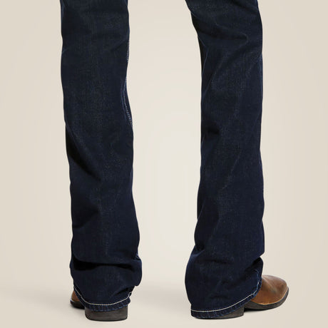 The image shows a person wearing the M7 Rocker Concord Stretch Stackable Straight Leg Jean - Dodge (10026681) in dark blue, paired with brown cowboy boots. The stylish jeans offer comfort with stretch fabric, as the individual stands with their back facing the camera against a plain light beige background.