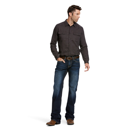 A man in a dark button-up shirt and M7 Rocker Concord Stretch Stackable Straight Leg Jeans stands on a white background, looking left. With one hand in his pocket, he complements his outfit with brown shoes.