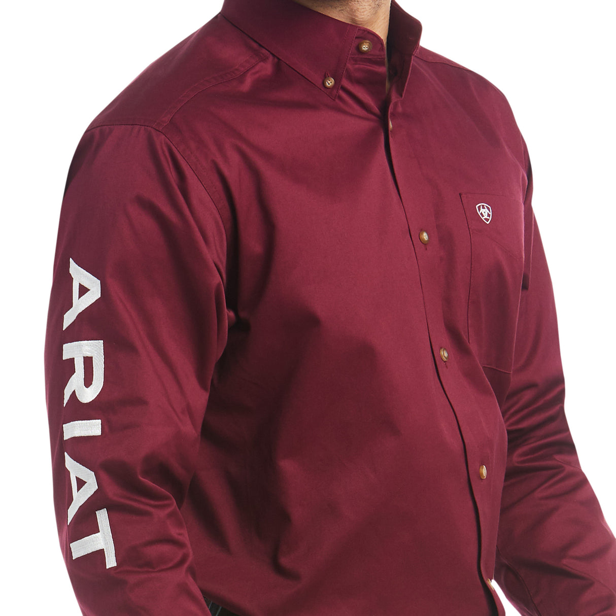 Wearing the Team Logo Twill Classic Fit Shirt Burgundy - 10027995, a person dons a premium cotton maroon button-up with a left chest pocket and shield logo. The right sleeve features ARIAT in bold white letters, creating a simple yet polished look.