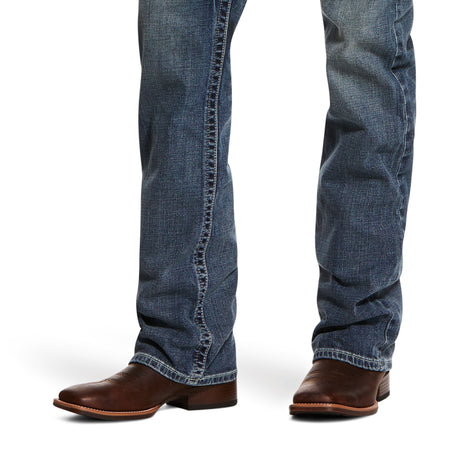 A pair of legs wearing M5 Slim Stretch Adkins Stackable Straight Leg Jean in lennox, with durable stitching and a slight fade, paired with pointed-toe, small-heeled brown leather cowboy boots against a plain white background.