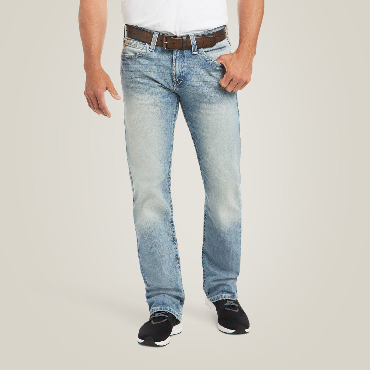 Someone in a white shirt, light blue M7 Rocker Stretch Stirling Stackable Straight Leg Jean - Shasta (10031997), and black shoes stands against a plain backdrop. The slightly faded jeans with stretch fabric are complemented by the brown belt held in one hand. The image is cropped at the shoulders.