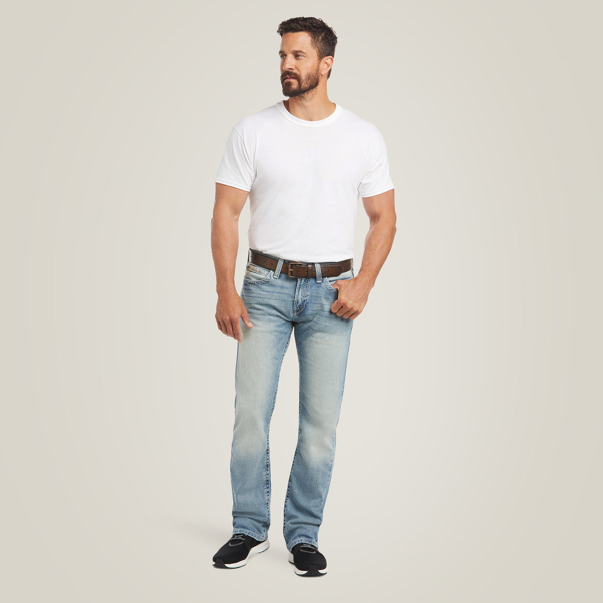 Against a plain background, a man with a beard and short hair wears a white T-shirt and light blue M7 Rocker Stretch Stirling Stackable Straight Leg Jean by Ariat in Shasta. He complements his outfit with a brown belt and black sneakers, looking to his left with his hands by his sides.