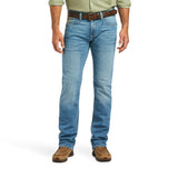 A modern cowboy exudes confidence in light blue M7 Slim Stretch Julian Straight Jeans, a brown leather belt, a green long-sleeve shirt, and tan suede shoes. Their arms rest casually at their sides against a plain white background.