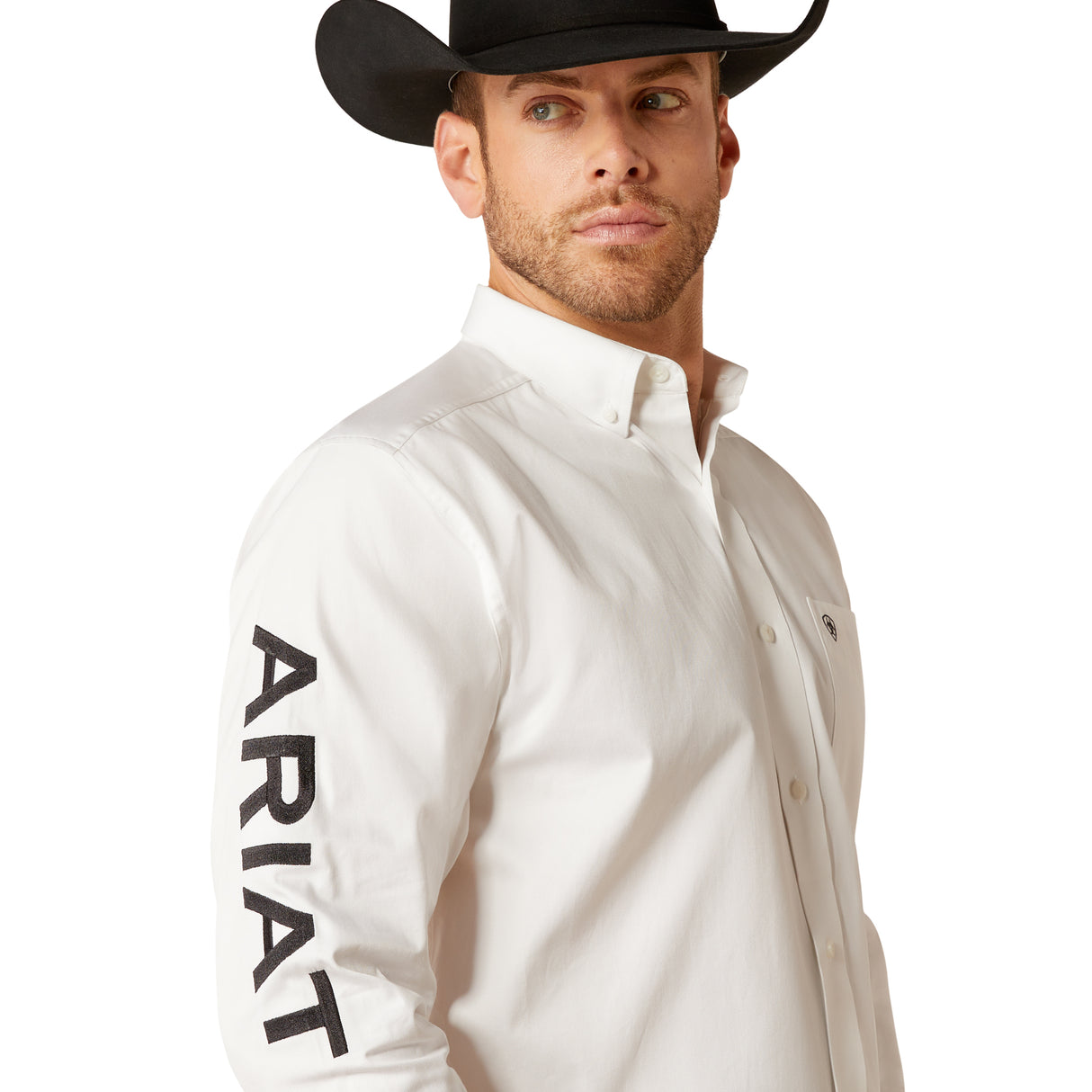 A man in a white ARIAT Team Logo Twill Classic Fit Shirt (10046825) glances left. His black cowboy hat pairs well with the subtle chest pocket logo and embroidered details, enhancing the timeless appeal of the shirts classic fit and plain white background.