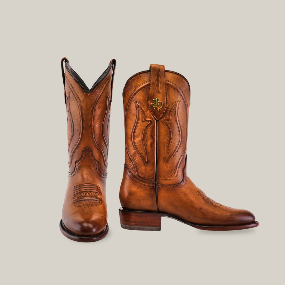 The Western Platinum Honey boots, in brown leather with intricate stitching and a decorative emblem on one boot, showcase classic cowboy style. They feature a roper toe and stacked heel, perfectly displayed against a plain white background.