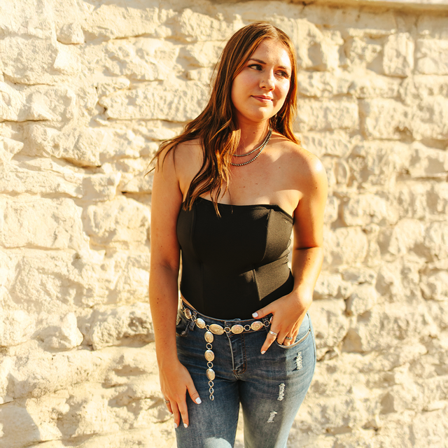 A woman with long brown hair stands against a beige stone wall, wearing the versatile LB Black Jumper Corset and distressed jeans. The corset-inspired top complements her thoughtful expression, one hand on her hip and the other by her side.